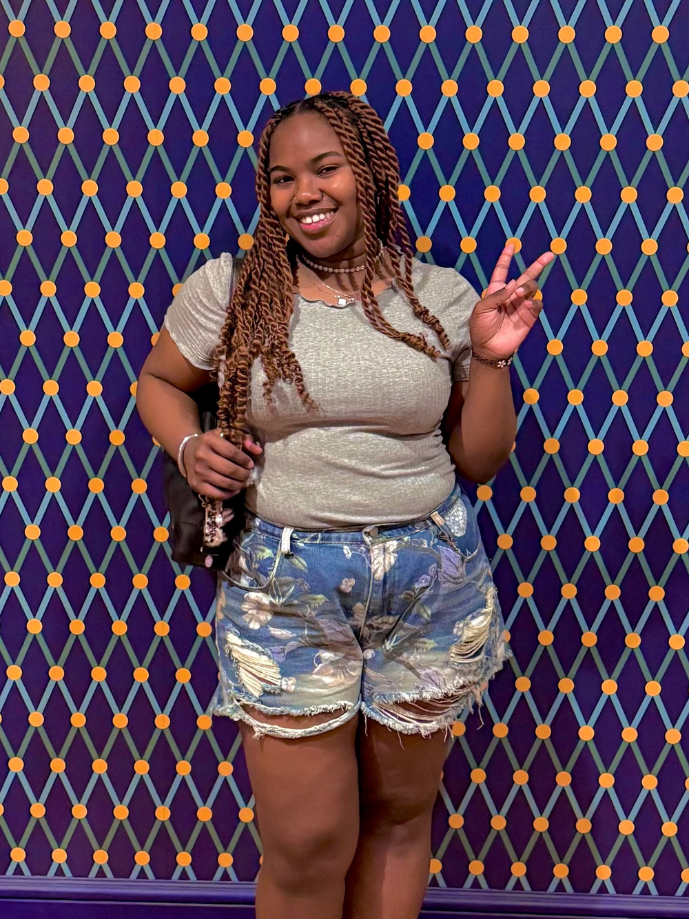 A woman posing in front of a blue and orange patterned wall, wearing a grey top, distressed denim shorts, and white sneakers.